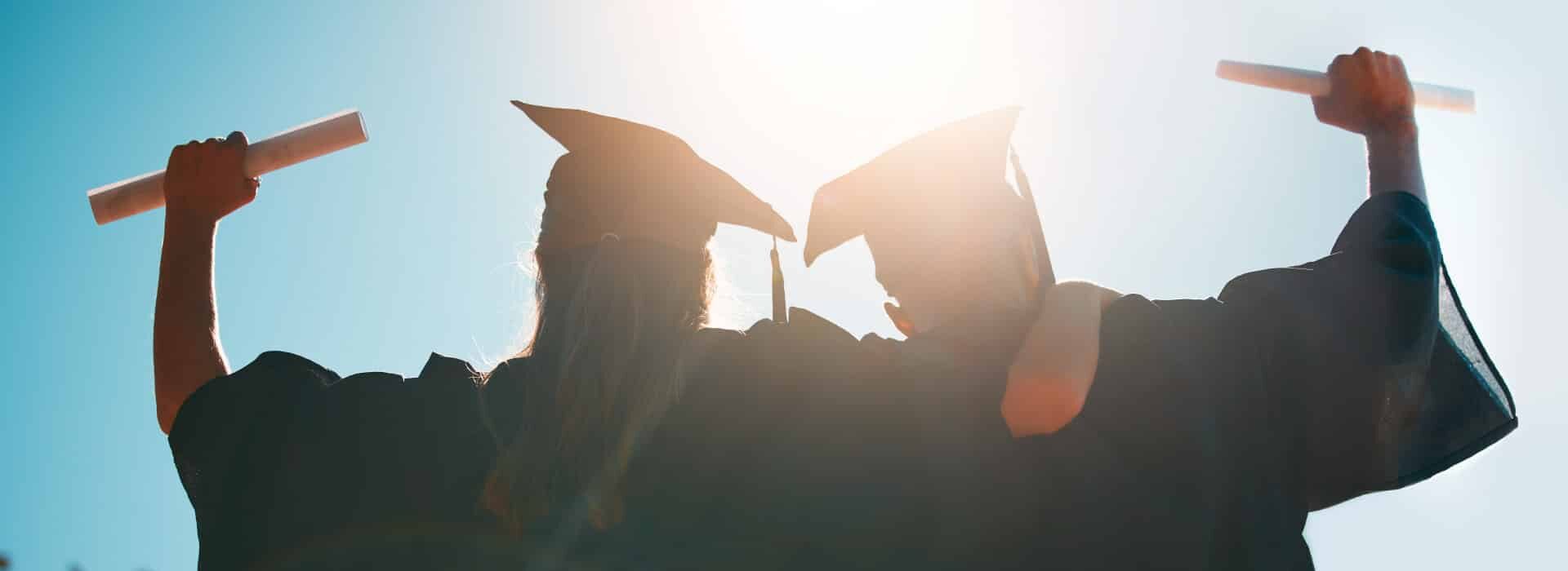 Rear view silhouette of university graduates in caps and gowns cheering and raising their diplomas toward the bright sunlight.