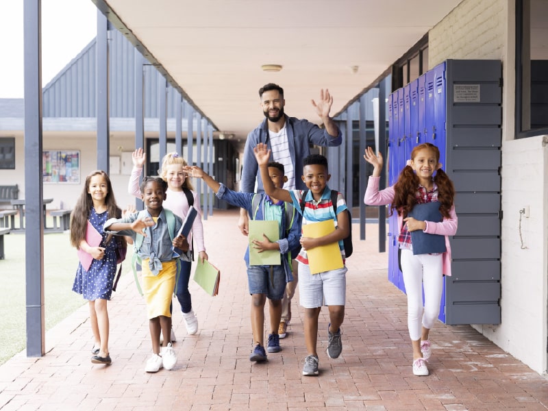 Diverse elementary school students and a teacher waving in a school entrance.