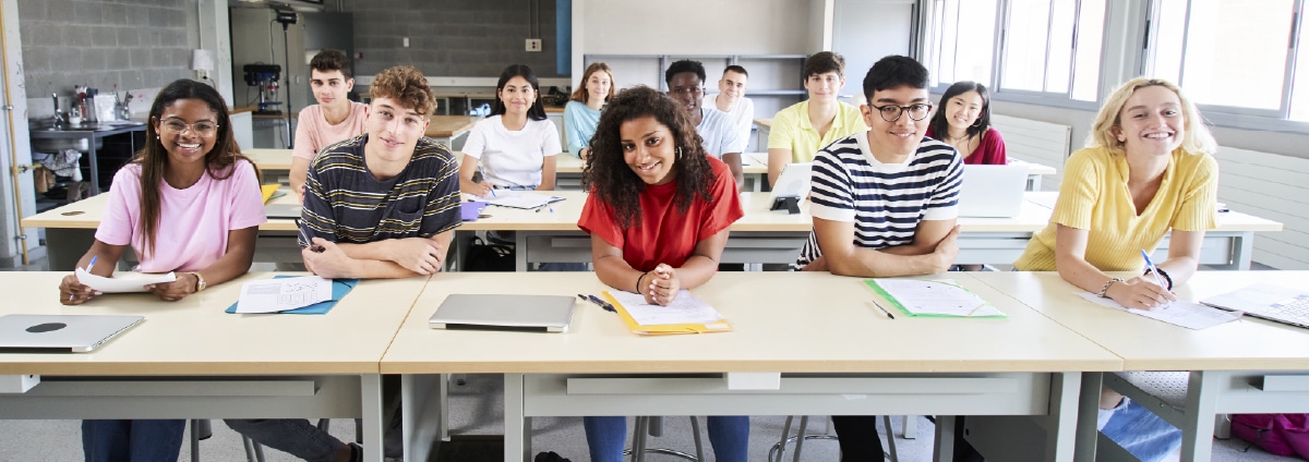 A diverse group of students sitting at desks in a brightly lit classroom, listening attentively to a lesson.